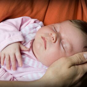 Ein schlafendes Baby liegt in einem Tuch. Eine Frau Hält ihre Hand schützend an den Kopf des Babys.